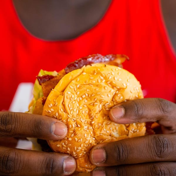 Hands holding a sesame seed bun burger with bacon and lettuce against a red background.