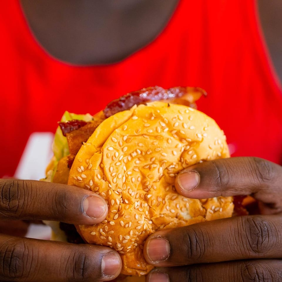 Hands holding a sesame seed bun burger with bacon and lettuce against a red background.