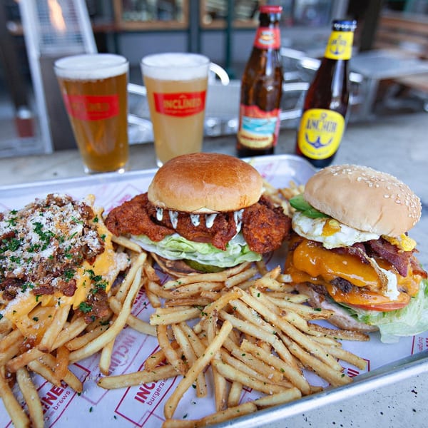 Inclined Burgers and Brews tray with two burgers, loaded fries, regular fries, and two beers.